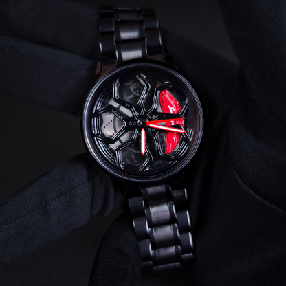 A black metal link Lamborghini Wheel Spinning watch with a glossy black dial, featuring a red butterfly-shaped hour marker and red hands, resting on a black fabric background.