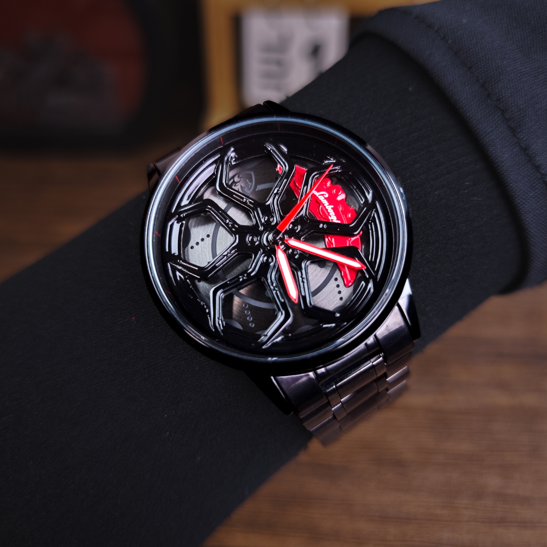 A close-up of a Lamborghini wristwatch with a black metallic case and a black dial featuring bold red hands and red accents, worn on a person's wrist with a black sleeve.