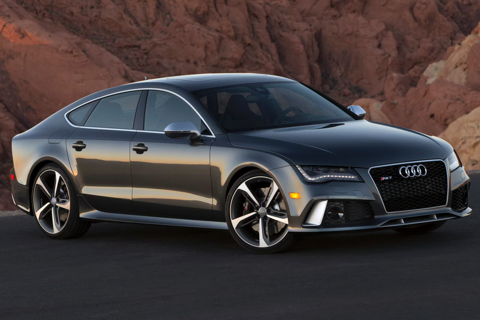 A dark gray metallic Audi S7 sports sedan parked outdoors against a backdrop of red rock desert mountains.